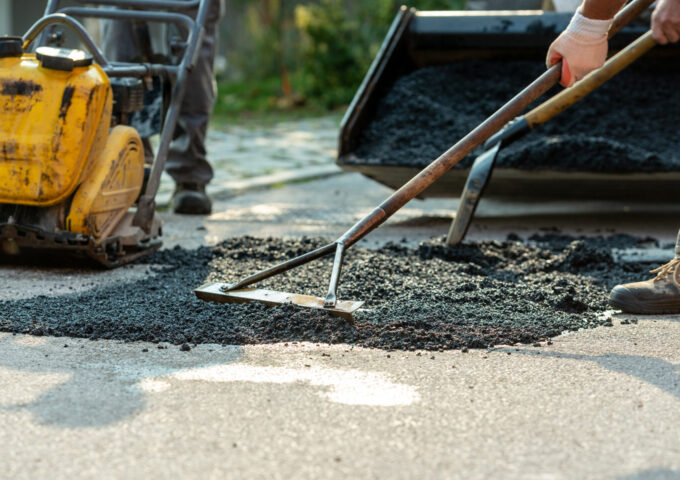 Low angle view of two workers arranging fresh asphalt mix with rakes and shovel to patch a bump in the road with machinery by their side.